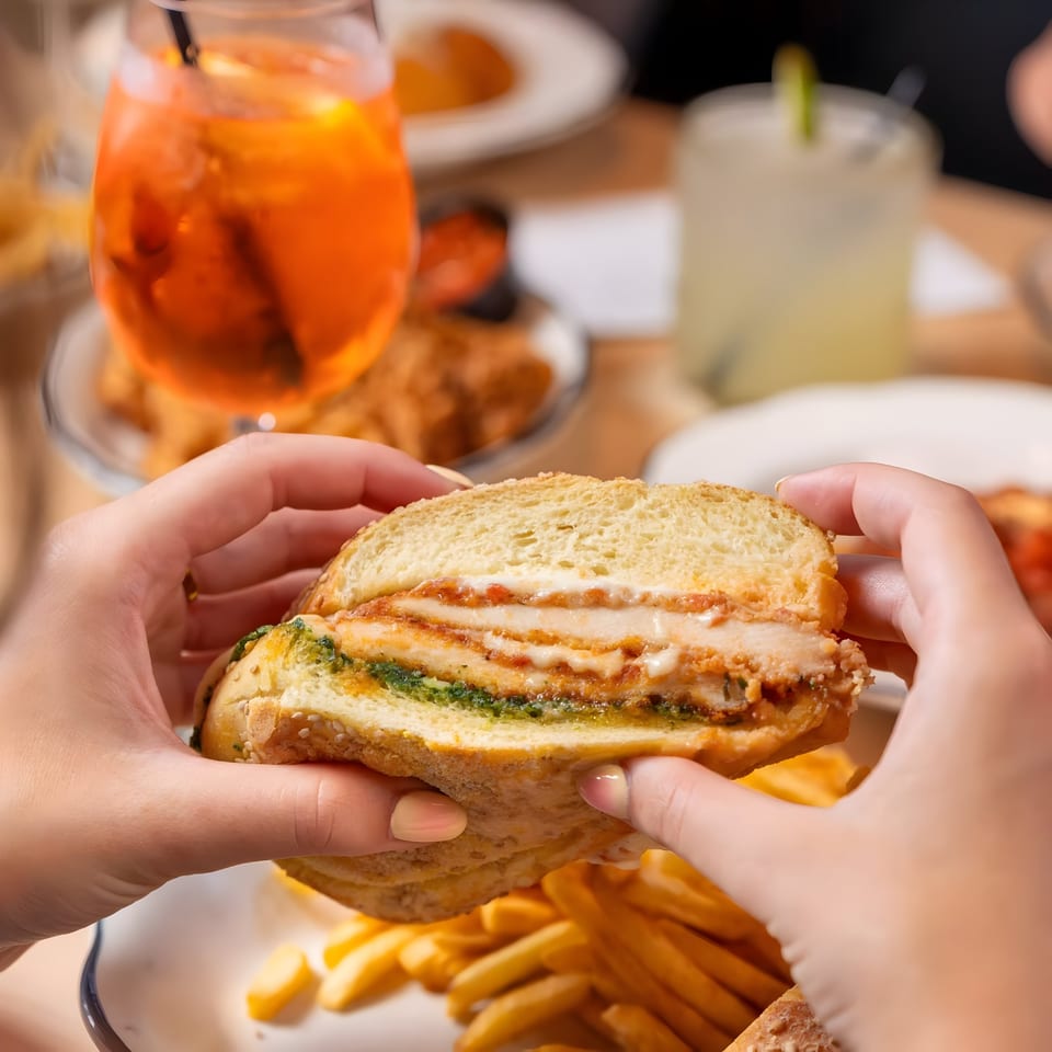 Hands holding a chicken sandwich with pesto and cheese, with fries and drinks in the background.