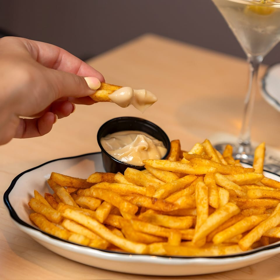 A hand dips a golden french fry into a small black cup of creamy sauce, with a martini in the background.